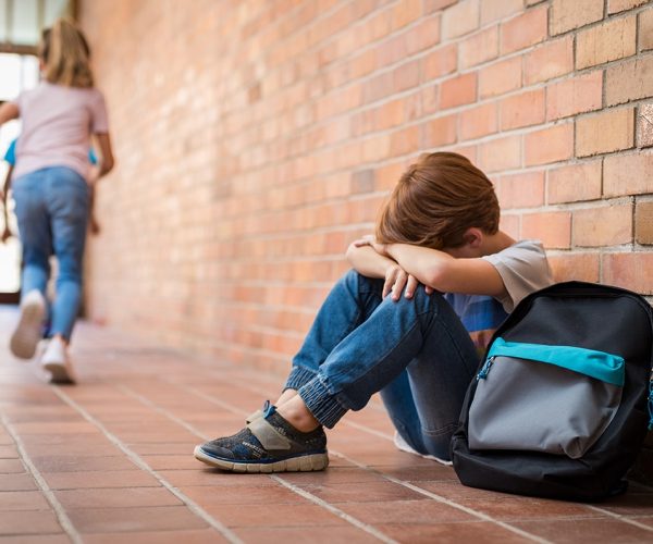 Little boy sitting alone on floor after suffering an act of bullying while children run in the background. Sad young schoolboy sitting on corridor with hands on knees and head between his legs.